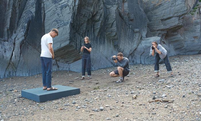 A model stands on a platform by a rocky cliff as three photographers capture images on a pebbled beach. A creative team in casual clothing surrounds t